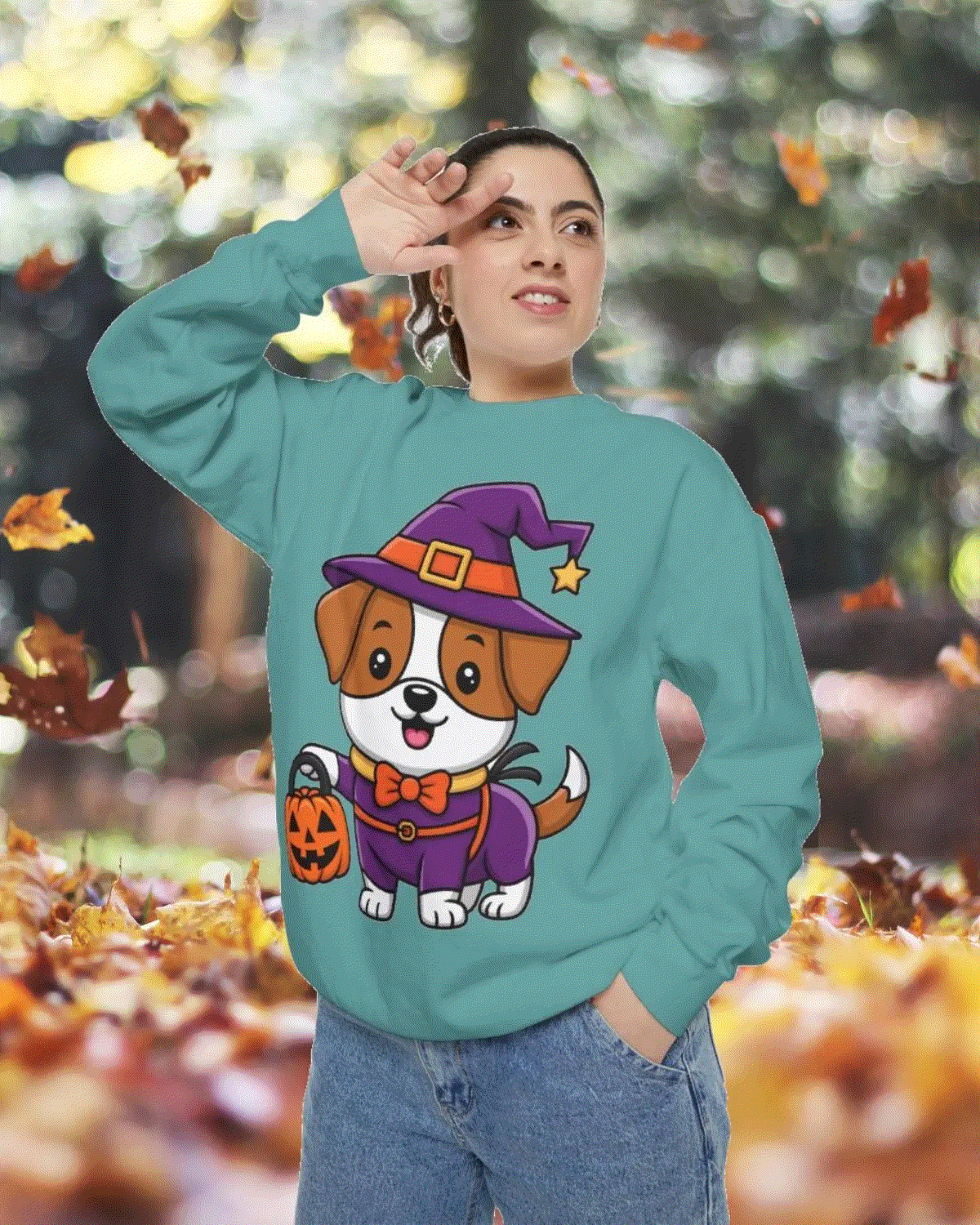 Person wearing teal Halloween sweatshirt with cartoon dog in witch costume holding pumpkin bucket, autumn leaves background