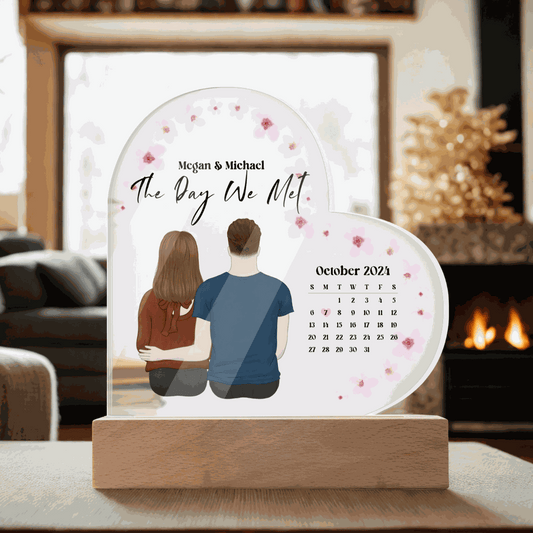 Personalized acrylic heart plaque with couple illustration, "The Day We Met" text, and October 2021 calendar on wooden base.