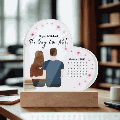Personalized acrylic heart plaque with couple illustration and calendar marking "The Day We Met" on wooden stand